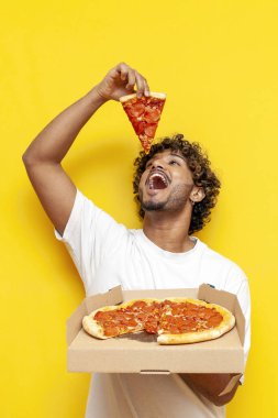Genç Hintli bir adam pizza kutusunu tutuyor ve sarı izole edilmiş arka planda bir parça yiyor. Kıvırcık öğrenci, ağzı açık ve dikey çerçeveli fast food gösterip reklamını yapıyor.