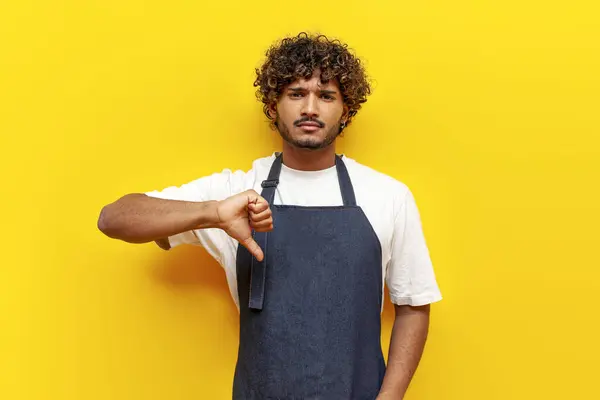 dissatisfied indian male waiter in apron shows dislike and bad rating on yellow isolated background, indian guy barista in uniform rejects and shows thumb down