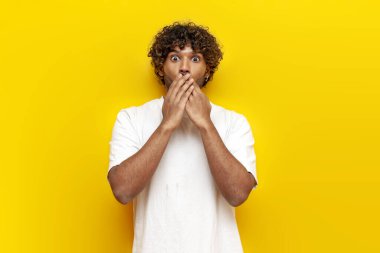 Şok olmuş beyaz tişörtlü genç Hintli adam şaşırır ve ellerini sarı izole arka planda tutar. Kıvırcık saçlı adam korku ve şaşkınlık içinde kameraya bakar.