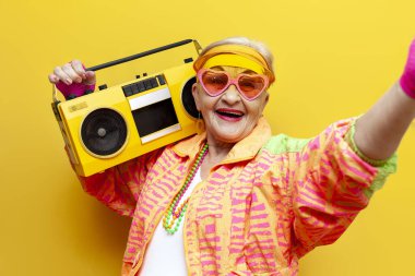 Hippi kıyafetleri içinde komik deli bir büyükanne teypten müzik dinliyor ve izole edilmiş sarı arka planda selfie çekiyor yaşlı havalı bir kadın partide dans ediyor ve gülümsüyor.