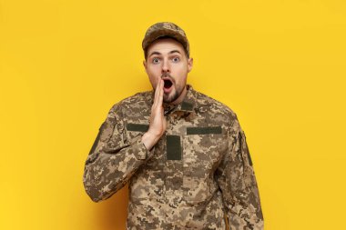 Kamuflaj piksel üniformalı Ukraynalı asker sarı izole arka plana şaşırmış görünüyor. Ukraynalı askeri öğrenci kameraya hayretle bakıyor.