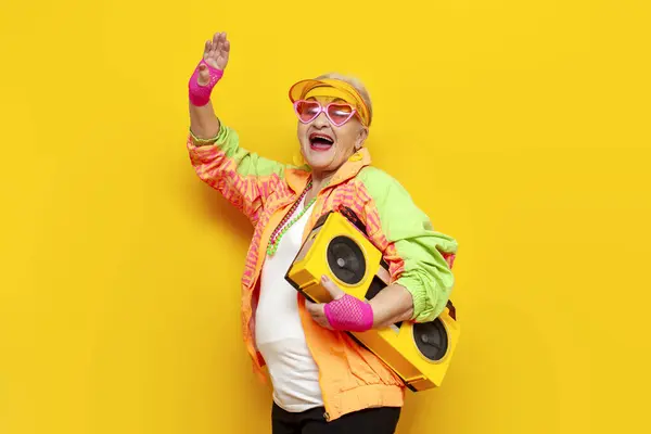 funny crazy granny in hipster clothes listening to music on tape recorder and singing on yellow isolated background, elderly cool woman dancing at party and screaming