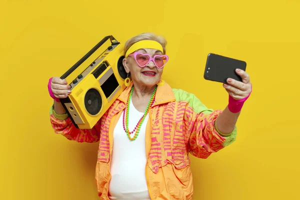 shocked crazy funny granny in hipster clothes listening to loud music on tape recorder online and using smartphone on yellow isolated background, surprised elderly woman pensioner holding mobile phone