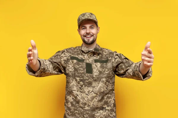 Ukrainian army soldier in pixel camouflage uniform hugs and raises his hands forward on a yellow isolated background, Ukrainian military cadet holds empty hands and shows