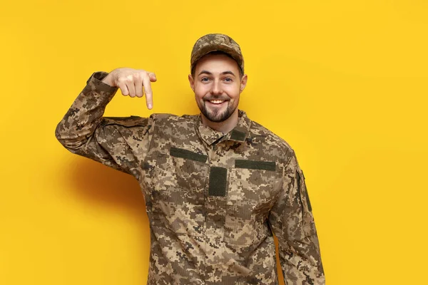 Ukrainian army soldier in camouflage uniform pixel pointing down on yellow isolated background, Ukrainian military cadet showing and advertising copy space from below