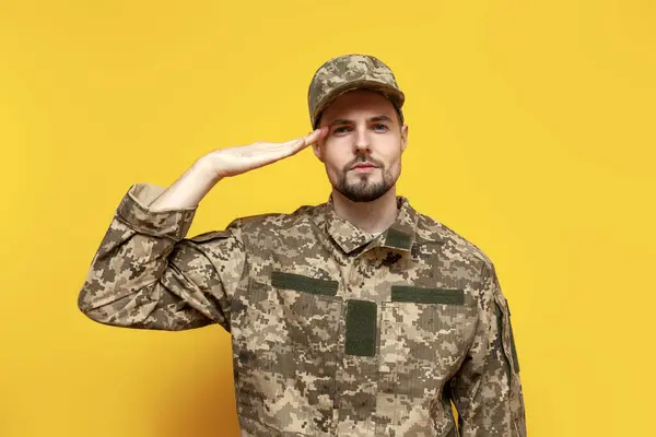 Ukrainian army soldier in pixel camouflage uniform salutes on yellow isolated background, Ukrainian military cadet shows attention gesture and executes command