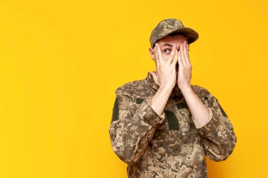 Genç Ukraynalı asker kamuflaj piksel üniforması içinde sarı izole bir arka planda saklanıyor ve dikizliyordu. Ukraynalı askeri öğrenci korkuyla yüzünü kapatıyordu.