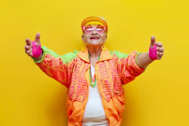 Hippi elbiseli, gözlüklü, şapka takan, sarı izole edilmiş arka planda selamlaşan, modaya uygun gençlik yıllarında elini boş tutan ve hoş geldin jestleri yapan yaşlı, havalı bir kadın.