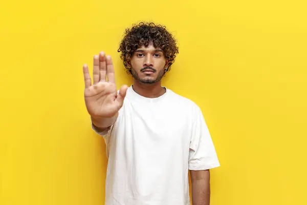 young indian man in white t-shirt prohibits and shows stop gesture on yellow isolated background, curly guy shows palm warns and refusal
