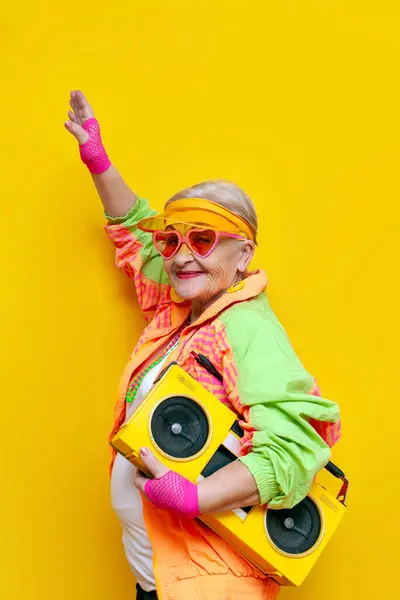 funny crazy granny in hipster clothes listening to music on a tape recorder and showing a kiss on a yellow isolated background, elderly cool woman dancing at a party and having fun