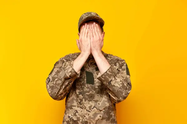 young Ukrainian army soldier in camouflage pixel uniform hiding and covering his face with his hands on a yellow isolated background, Ukrainian military cadet is shy and remains unrecognizable