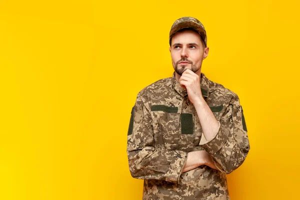 pensive young Ukrainian army soldier in camouflage uniform pixel plans and thinks on a yellow isolated background, Ukrainian military cadet dreams and imagines
