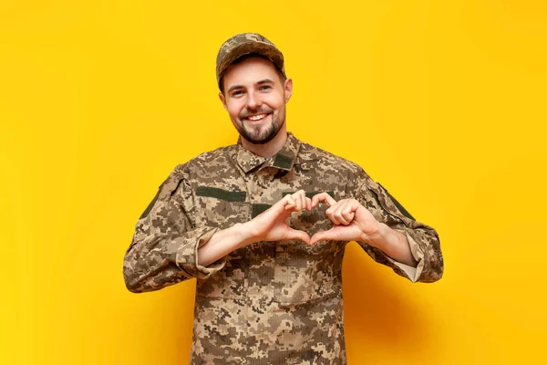 young Ukrainian army soldier in pixel camouflage uniform shows heart with hands on yellow isolated background, Ukrainian military cadet shows gesture of love and charity