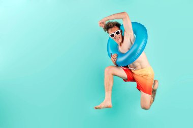 Şişirilebilir yüzme yüzüğüyle kazanan genç adam yazın mavi arka planda atlıyor ve uçuyor. Erkek turist plajda dinlenmek için koşuyor ve zaferle seviniyor.