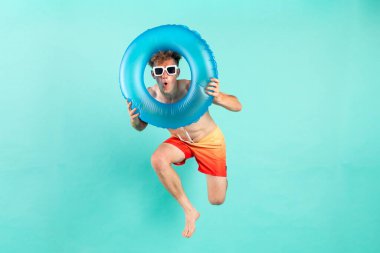 Şişirilebilir yüzme yüzüğüyle kazanan genç adam yazın mavi arka planda atlıyor ve uçuyor. Erkek turist plajda dinlenmek için koşuyor ve zaferle seviniyor.