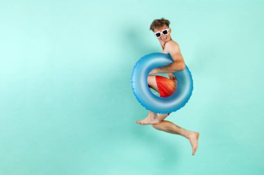 Şişirilebilir yüzme yüzüğüyle kazanan genç adam yazın mavi arka planda atlıyor ve uçuyor. Erkek turist plajda dinlenmek için koşuyor ve zaferle seviniyor.