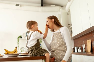 Önlüklü genç anne, evdeki mutfakta yemesi için küçük oğluna kurabiye veriyor, 2 yaşındaki çocuk ailesiyle kahvaltı ediyor, kadın çocuğu besliyor.