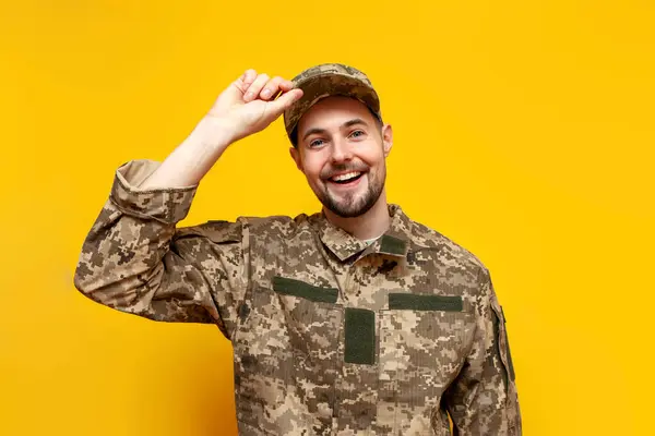 young Ukrainian army soldier in camouflage pixel uniform smiling on yellow isolated background, Ukrainian military cadet putting on cap