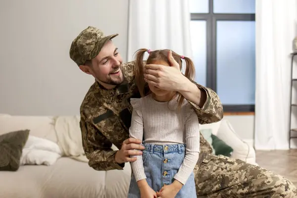 happy soldier of Ukrainian army in camouflage uniform returned home and meets his daughter, military dad surprises the child and closes his eyes