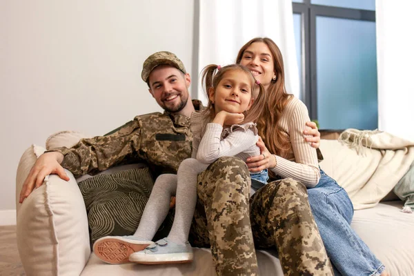 Ukrainian military man in camouflage uniform hugs his family at home and smiles, a happy soldier of the Ukrainian army returned home and sits on the sofa with his wife and daughter, mobilization