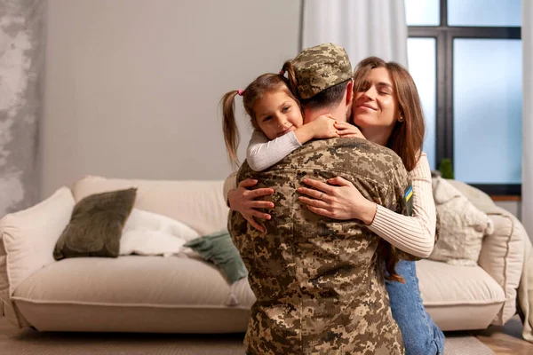 Ukrainian military man in camouflage uniform hugs his family at home and smiles, a happy soldier of the Ukrainian army returned home and sits on the sofa with his wife and daughter, mobilization