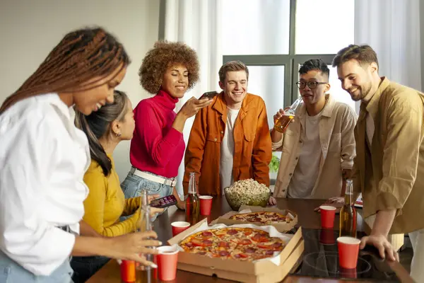 multiracial group of young people at house party ordered pizza and beer and photographed food on phone, students of different ethnicities at house party drink alcohol and eat fast food and have fun