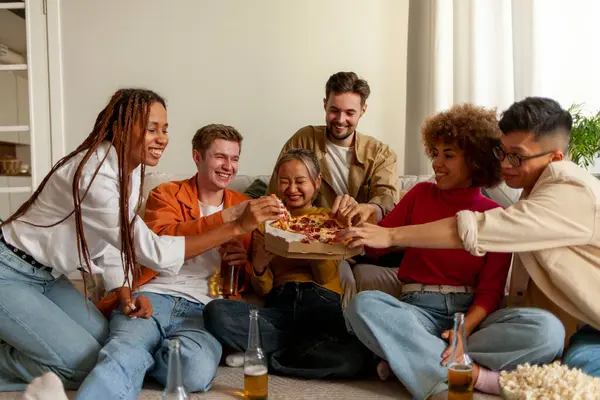 multiracial group of young people at house party eating pizza and drinking beer and having fun with friends, students of different ethnicities drinking alcohol and eating fast food and celebrating