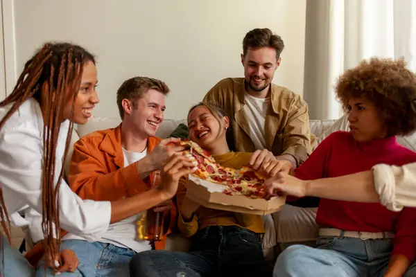 multiracial group of young people at house party eating pizza and drinking beer and having fun with friends, students of different ethnicities drinking alcohol and eating fast food and celebrating