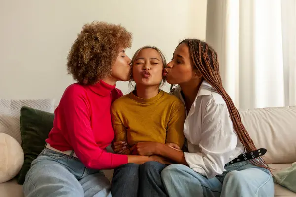 African American women and Asian woman sitting on sofa at home and hugging, interracial girlfriends smiling and kissing