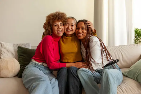 African American women and Asian woman sitting on sofa at home and hugging, interracial girlfriends smiling and looking at camera