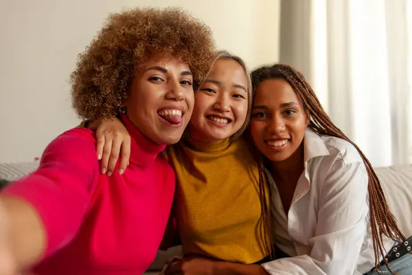 African American women and Asian woman sitting on sofa at home taking selfie and hugging, interracial girlfriends smiling and showing tongue and taking photos on phone