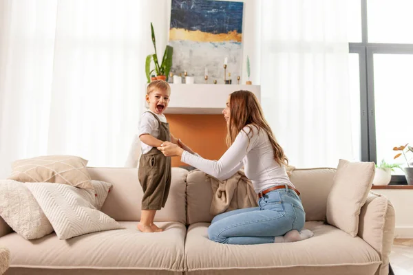 young mother and her little son dance and jump on the sofa at home, 2-year-old boy screams and rejoices with his parent, woman has fun with her child