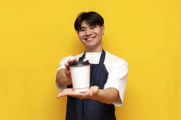 asian young male waiter in apron giving cup of coffee on yellow isolated background, korean guy barista businessman in uniform holding drink in paper cup