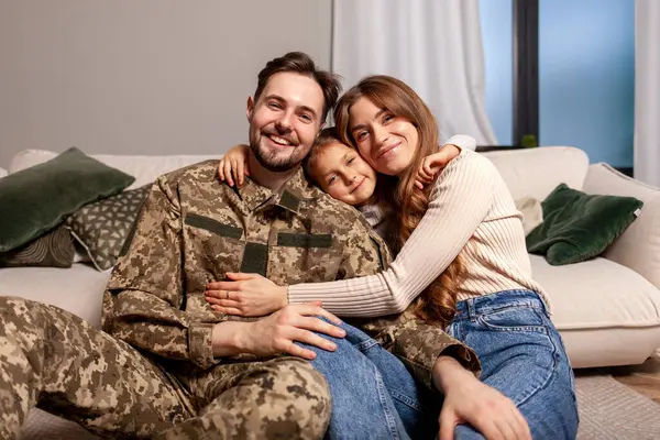 Ukrainian army soldier in camouflage uniform returned home to his family, military cadet sits on the sofa with his wife and daughter and smiles, child hugs veterans father
