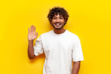Beyaz tişörtlü neşeli Hintli genç adam sarı yalıtılmış arka planda el sallıyor, kıvırcık adam el hareketi yapıyor ve elveda diyor.