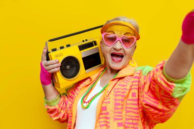 Hippi kıyafetleri içinde komik deli bir büyükanne teypten müzik dinliyor ve izole edilmiş sarı arka planda selfie çekiyor yaşlı havalı bir kadın partide dans ediyor ve gülümsüyor.