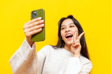 Genç mutlu Asyalı kadın diş telleriyle selfie çekiyor ve sarı izole edilmiş arka planda barış jesti gösteriyor, Koreli kız görüntülü sohbet ediyor ve selamlıyor.