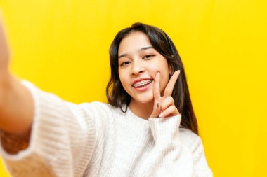 Genç mutlu Asyalı kadın diş telleriyle selfie çekiyor ve sarı izole edilmiş arka planda barış jesti gösteriyor, Koreli kız görüntülü sohbet ediyor ve selamlıyor.