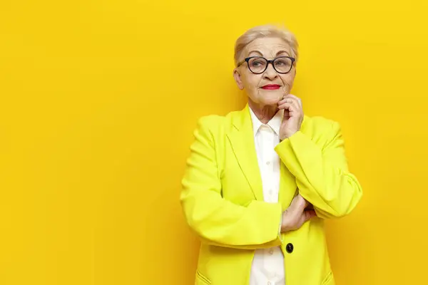 pensive old businesswoman in glasses and business clothes planning and thinking on yellow isolated background, elderly pensioner grandmother in blazer dreaming and imagining