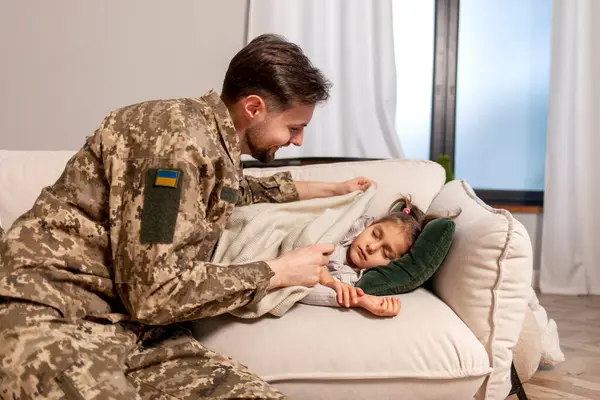 happy soldier of Ukrainian army in camouflage uniform returned home and puts his daughter to bed, military dad protects and takes care of child at home, concept of mobilization in Ukraine
