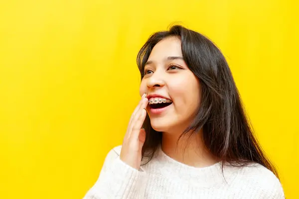 young modest asian girl with braces smiling and taunting on yellow isolated background, korean girl covering her smile and being shy