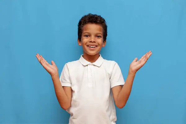 stock image african american teenage boy in white polo smiling and holding empty hands on blue isolated background, cheerful child showing copy space and empty palms