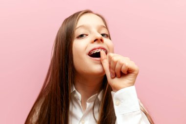 Bahse girerim öyledir. Diş teli takan, umut verici hareketler gösteren, pembe arka planda parmağı olan, dişlerine dokunan, diş tebessümü ve diş bakımı gösteren küçük bir kız çocuğu.