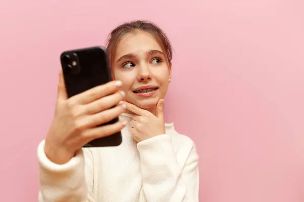 unsure teenage girl with braces uses a smartphone and plans on a pink isolated background, a thoughtful child with a mobile chooses and doubts