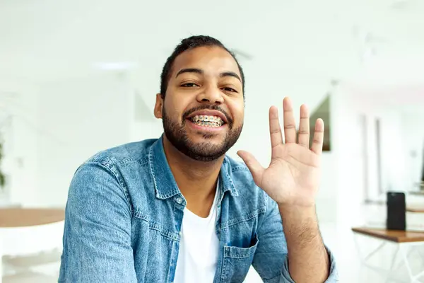 happy african american man with braces greets and waves in white cafe, man in denim shirt shows palm and hi and bye gesture