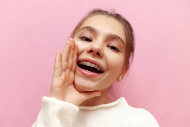 Diş teli takmış neşeli bir genç kız pembe izole bir arka planda bilgi veriyor, çocuk ağzı açık konuşuyor.