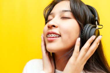 Telli genç Asyalı kadın kulaklıkla müzik dinliyor ve sarı izole arka planda şarkı söylüyor, Koreli kız müzikle dans ediyor, yakın plan.