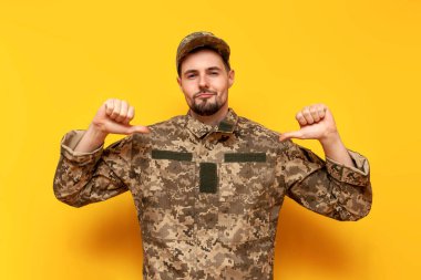 Piksel kamuflaj üniformalı gururlu genç Ukraynalı asker ellerini sarı izole edilmiş bir geçmişe doğrultuyor. Ukraynalı askeri öğrenci kendini gösteriyor ve reklamını yapıyor.