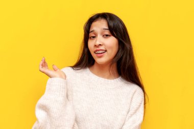 Genç, kendine güveni olmayan Asyalı kız omuzlarını sarı yalıtılmış arka planda silkeliyor, kafası karışmış Koreli kız kayıtsızlık gösteriyor ve elini kaldırıyor.