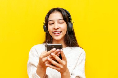Telli genç Asyalı kadın akıllı telefon kullanıyor ve kulaklıklı müzik dinliyor sarı izole arka planda, Koreli kız telefonla gülümsüyor.